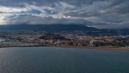 Fototapeta premium Aerial drone view of Dénia on the Mediterranean coast of Spain with Montgó mountain and dramatic storm clouds. The historic fortress hill rises above dense urban architecture and coastal landscape.