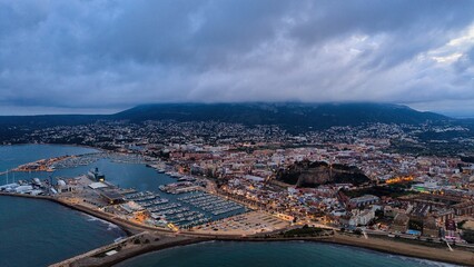 Obraz premium Aerial drone view of Dénia harbor and Mediterranean coastline with Montgó mountain under dramatic cloud formations. The marina, breakwater and coastal city stretch along the reflective sea.