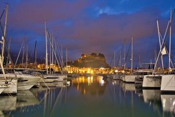 Obraz premium Marina harbor in Dénia on the Mediterranean coast of Spain filled with sailboats and yachts. The historic castle hill rises above the waterfront beneath dramatic sunset clouds reflected in calm water.