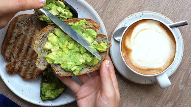 Top down view of female hands spreading fresh chopped avocado on a slice of toasted whole wheat bread with a knife, next to a cup of creamy cappuccino for a healthy and delicious breakfast