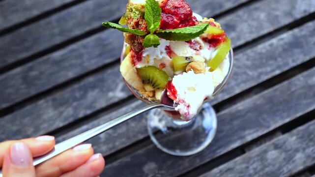 Woman's hand dipping a spoon into a delicious fruit parfait with cream, kiwi, raspberry, and mint, scooping a portion to eat from a tall glass on a dark wooden table outdoors