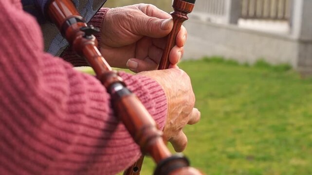 Close-up of an elderly man's hands carefully adjusting or assembling the wooden drone of a bagpipe in an outdoor setting.