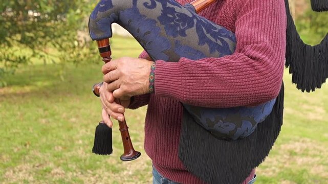 A medium shot of an older man playing a bagpipe with a blue floral pattern bag and black fringes, focusing on the coordination between bag and chanter.