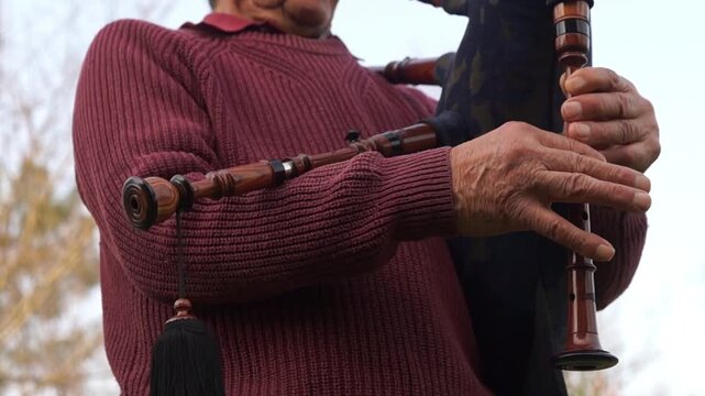 A low angle perspective showing the intricate finger movements of an elderly man on a wooden bagpipe chanter against a soft outdoor background.