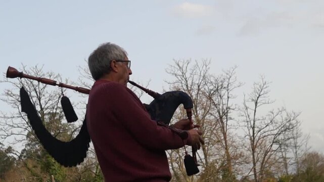 Wide shot of a senior man standing in a park, wearing a burgundy sweater, playing the bagpipes with trees and a cloudy sky in the background.