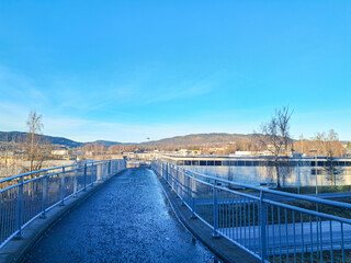 Pedestrian bridge walkway with metal railings leading toward town buildings and distant mountains under clear blue sky with copy space.