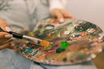 Young woman artist mixing paint on palette while painting on canvas at easel in art studio, creative hobby and painting process