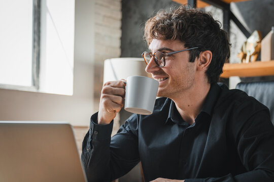 Businessman working on laptop at office desk, drinking coffee, smiling and thinking. Pleased young Caucasian man holding cup with hot beverage and looking through a window. Break from work