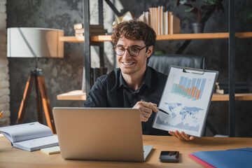 Fototapeta premium Man, video call laptop in office for paperwork, remote work and graphs for report. Freelancer, business documents and tech for project management, finance agency, giving consultation, conference app