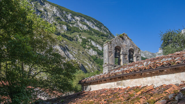 Detalle de una antigua espada&ntilde;a con dos campanas sobre un tejado de teja &aacute;rabe, rodeada de frondosos &aacute;rboles y las imponentes laderas rocosas de las monta&ntilde;as asturianas bajo un cielo azul.