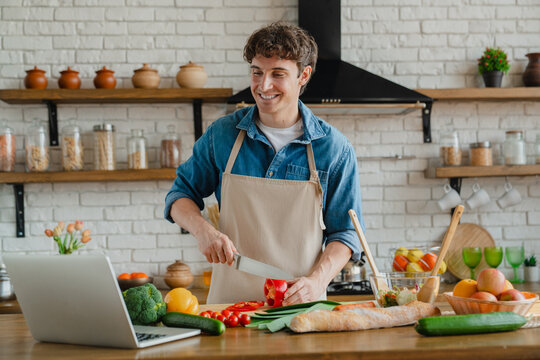 Smiling young Caucasian male chef in apron preparing salad in modern kitchen interior with organic vegetables and laptop. Food blog at home, new recipe to cook healthy dinner at home and diet