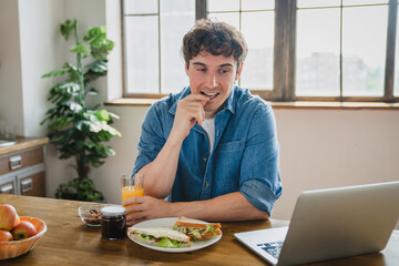 Smiling young man eating sandwich breakfast lunch using laptop work study remotely watching video...