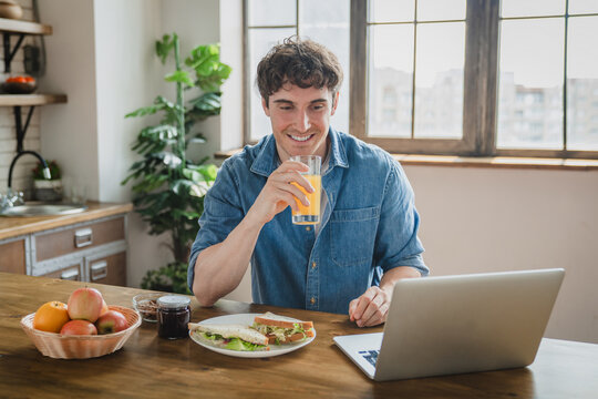 Laptop, breakfast and kitchen with man, sitting while browsing the internet. Computer, social media and orange fresh juice with young male freelance working study from home and morning eating