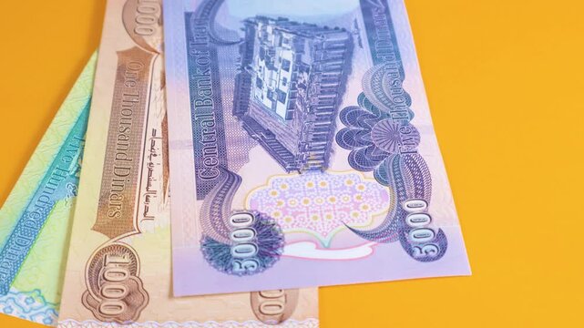 A hand places various Iraqi Dinar banknotes on a yellow background. The video displays denominations from 250 up to 50,000, highlighting the colorful designs and historical motifs of the currency.