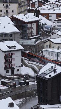 Aerial view of Zermatt, Switzerland, snow topped chalets, Le Chalet and Felsenheim, glass towers, cog train passes, cyclist and pedestrians move on wet curved streets.