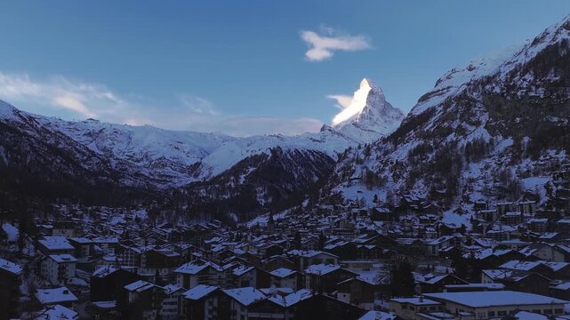 Aerial view of Zermatt, Switzerland, in winter beneath Matterhorn. Drone sweep shows chalets, ski lifts, rocky ridges, cool shadow, bright horn at sunrise or sunset.