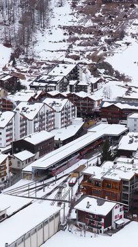 Aerial scene in Zermatt, Switzerland shows red mountain trains arriving and departing at the railway station, with chalet buildings and snow under overcast daylight.