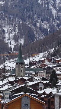 Aerial pan over Zermatt, Switzerland, with snow covered chalets, stone church tower with green spire and clocks, Zermatterhof sign, cranes, overcast winter light.