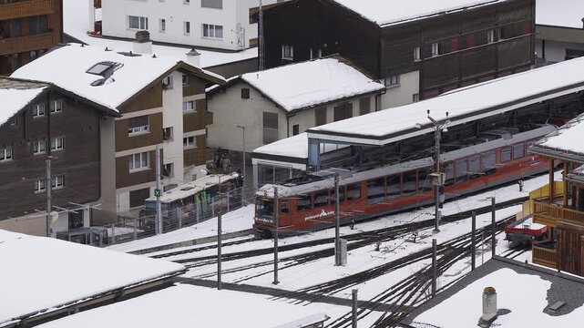 Aerial view shows orange Gornergrat Bahn cog train at covered platform in Zermatt, Switzerland, with snow, rack and pinion tracks, switches, chalets, and people waiting.