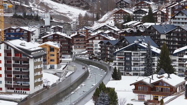 Aerial daylight pan of Zermatt, Switzerland shows chalets and flats on a steep hill, a channelized stream, a curving road, a service vehicle, and one pedestrian.
