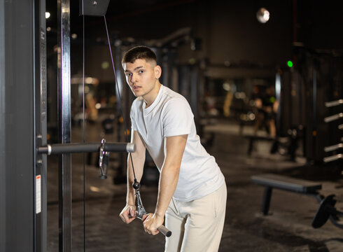 Focused young guy in white sportswear training arms on cable machine at gym, performing triceps pushdowns as part of workout routine