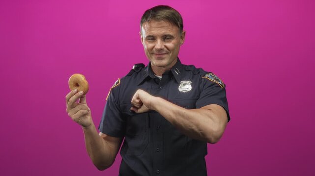Man police officer smiling and holding donut in left hand with right fist near chest in studio; pride humor.