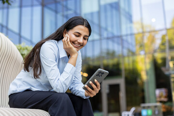 Fototapeta premium Businesswoman sitting outside a modern office building. Smiling and laughing while using her smartphone and wireless earbuds. Conveying connectivity. Digital communication. And relaxed professionalism