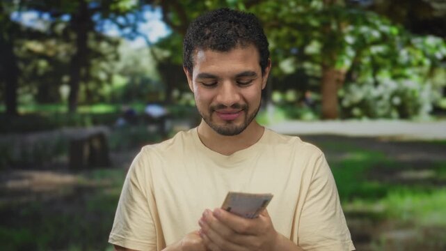 Casually dressed smiling young man fanning saudi riyal banknotes and counting money in a sunlit green park; happiness.