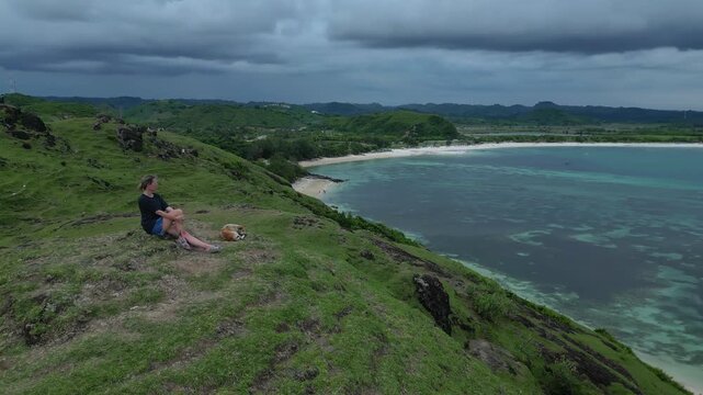 Woman Sitting Cliff Overlooking Turquoise Bay, Panoramic Aerial Viewpoint Showing Coral Reef Patterns, Distant Green Hills And Crescent Beach,Bukit Merese, Lombok, Tanjung Ann