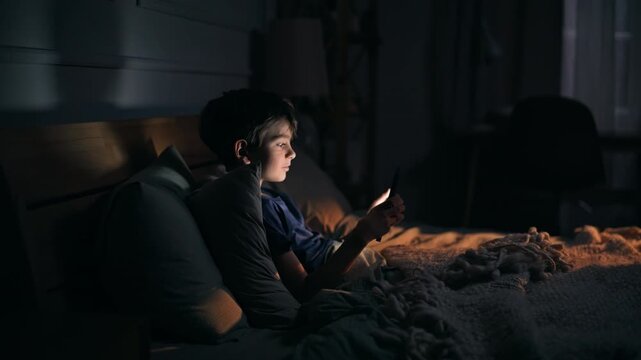 Boy playing smartphone bedtime illuminated screen glow in night bedroom closeup.