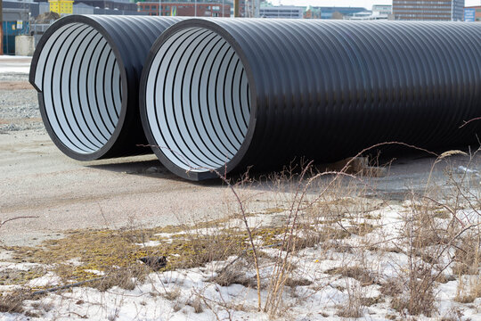 Large black drainage pipes with white interior stripes placed on a construction site, surrounded by gravel and sparse vegetation under a clear blue sky