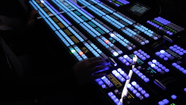 Close-up of hands operating a complex video switcher console in a dark broadcasting studio. The illuminated blue and purple buttons highlight the intricate controls used for live production.