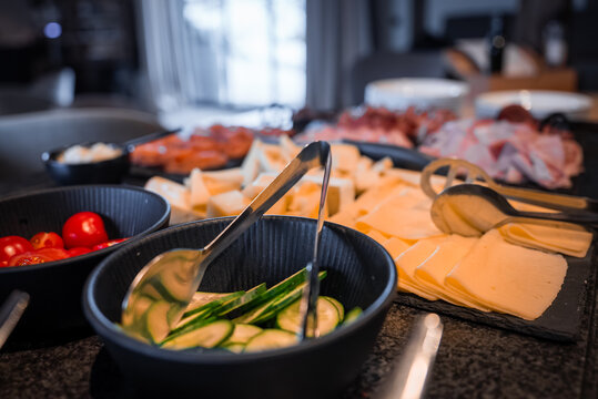 Neatly stacked semi hard cheese, cucumber, cherry tomatoes, and cured meats sit on dark stoneware with metal tongs in a boutique hotel in the Dolomites.