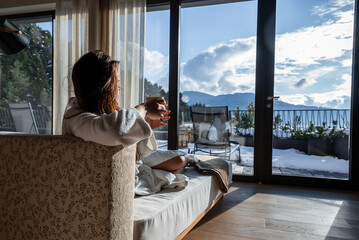 Fototapeta premium A guest lounges on a sofa with a cup, facing the Dolomites through tall windows. Snow dusts terraces and pine mountains. Private balcony and winter light set a calm mood.