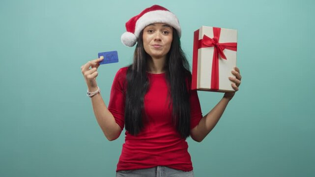 Woman holding a creditcard and a large giftbox smiles while wearing a santa hat in a studio; joyful holiday gifting.