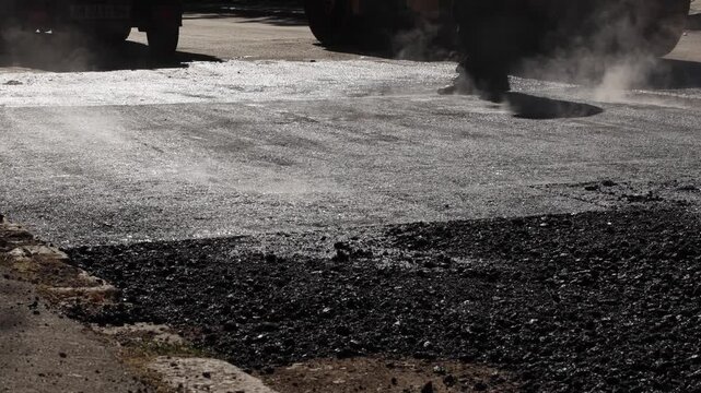 Close up of freshly laid hot asphalt surface during street construction. Steam rising from new pavement while road workers move around the construction area.