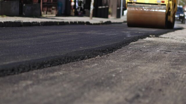 Road roller compacting fresh asphalt during street construction while cars drive past on nearby traffic lane in urban environment.