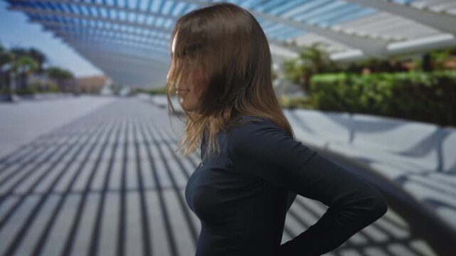 Young hispanic woman with hair over face holding lower back for backache on street under slatted canopy and long shadow patterns; mild discomfort.