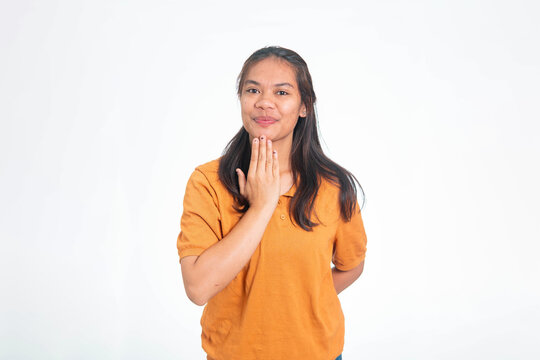 Young Indonesian woman in orange shirt using sign language to say thank you, smiling and gesturing with her hand near her chin.
