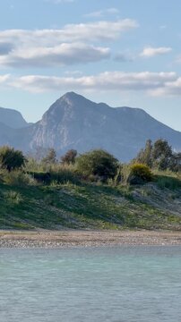 Small white caravan parked on a grassy slope near pebble beach. Calm water in the foreground and massive mountains in background. Green bushes and dry grass. A mobile home in nature, van life movement