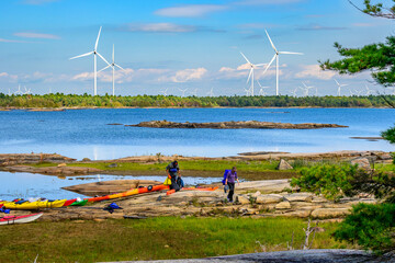Kayakers move equipment and bags across a rocky coast near parked boats with wind turbines in the...