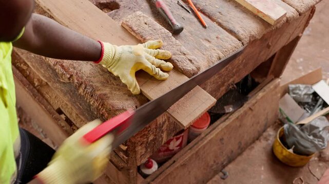 Over-the-shoulder shot of local African male carpenter in work gloves sawing a wooden plank with a hand saw on a rustic workbench in his workshop