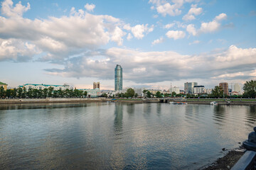 Obraz premium Modern buildings and green areas along the iset river in yekaterinburg, russia, create a picturesque cityscape under a cloudy sky