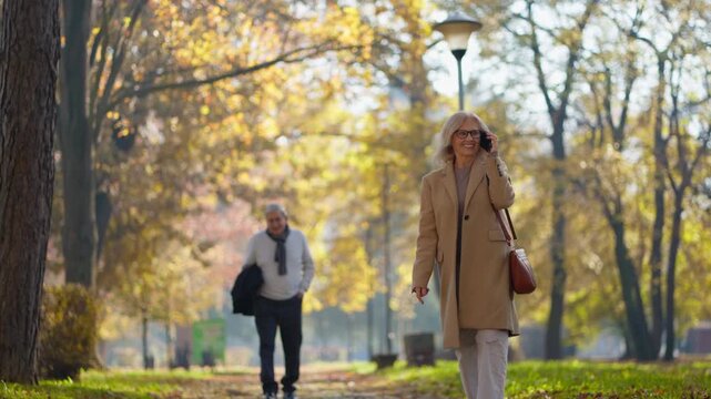 Smiling woman in a coat talking on her phone while walking in a park during autumn