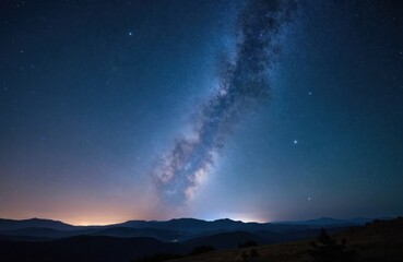 Fototapeta premium Night sky over Portugal mountains shows Milky Way galaxy. Stars shine bright in dark blue expanse. Lights glow on distant horizon from small town.