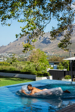 Mature African man lying and paddling on clear float in home infinity pool wearing peach trunks