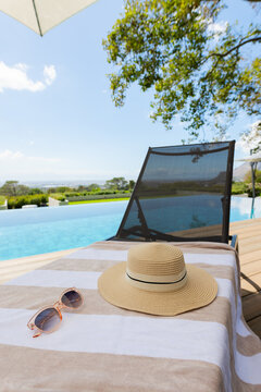 Woven straw sun hat sitting on striped towel on wooden deck, with sunglasses and infinity pool