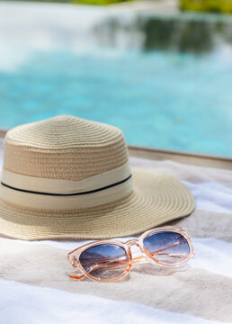 Straw sun hat and transparent sunglasses are resting on striped towel poolside, sunlight reflecting