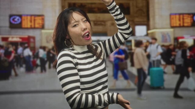 Woman singing with hands raised in a busy train station concourse among rushing travelers and rolling suitcases, striped sweater visible; joyful energy.