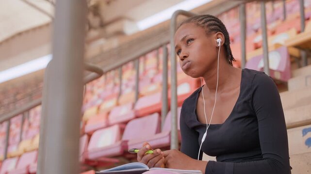 black student listening with earphones, gazing upward between note taking, thoughtful expression and braided hair, stadium seating background and afternoon light, introspective creative planning